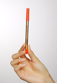 KIKO krämig färgpenna, med ett guldigt hölje och en klar orange kork och spets, hålls mellan fingrarna mot en neutral bakgrund.