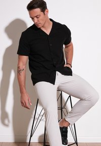 Camisa negra de manga corta con botones y cuello plano, combinada con pantalones ajustados gris claro y zapatos negros tipo slip-on, sentado en un taburete de alambre negro.