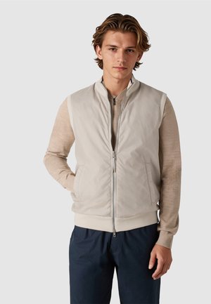 Ung mand iført en lys beige lynlåsvest over en beige langærmet skjorte og mørkeblå bukser, stående med den ene hånd i lommen.