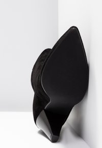 Czarne zamszowe buty na wysokim obcasie z wyprofilowanym noskiem i teksturowaną czarną gumową podeszwą, ustawione pod kątem na białym tle.