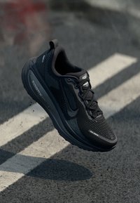 Svart Nike joggesko i luften over en asfaltert vei med hvite linjer, som viser detaljer av sålen, mesh-overdelen og snøringen.