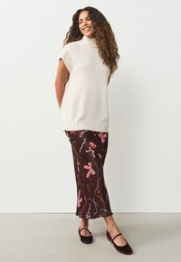 Pull sans manches col roulé beige clair, associé à une jupe midi marron à motifs avec des dessins floraux roses et abstraits ; chaussures bordeaux.
