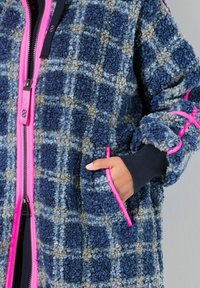 Cappotto oversized in tessuto a scacchi blu e navy con zip e rifiniture rosa, dotato di tasche laterali e dettagli a coste sui polsini. Texture morbida.