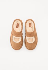 Tan suede klippere med en myk, kremfarget shearling-fôr, med lukket tå og en bred stropp over foten for en sikker passform.