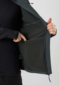 Person iført sort tøj viser indersiden af en mørkegrøn jakke med lynlås, lavet af tekstureret stof og med en inderlomme.