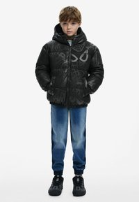 Black puffer jacket with Spider-Man graphics, zip front, and hood. Dark blue jeans, elastic cuffs, and black sneakers complete the outfit.