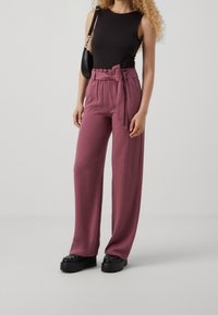 Pantalon violet taille haute avec ceinture nouée, fabriqué dans un tissu doux, associé à un haut noir sans manches et des chaussures noires épaisses.