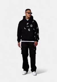Sweatshirt à capuche noire avec des graphismes de phases de lune, associé à un pantalon cargo noir. Comprend un t-shirt blanc et des baskets blanches.
