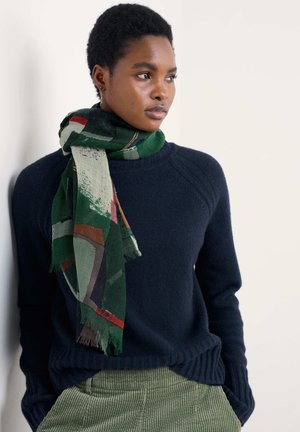 Multifarvet tørklæde i grøn med abstrakte mønstre og frynset kant, båret over en marineblå sweater med ribbet halsudskæring og grønne fløjlsbukser.