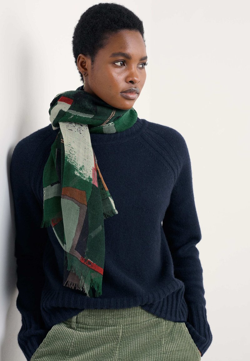 Multifarvet tørklæde i grøn med abstrakte mønstre og frynset kant, båret over en marineblå sweater med ribbet halsudskæring og grønne fløjlsbukser.