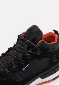 Gros plan sur une basket noire avec doublure orange, lacets noirs à pois orange et logo Timberland visible sur la languette et le côté.