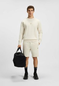 Lichtbeige sweatshirt en shorts, gestructureerde stof, met een logo patch. Zwarte duffel tas, zwarte sokken met witte branding en zwarte sneakers.