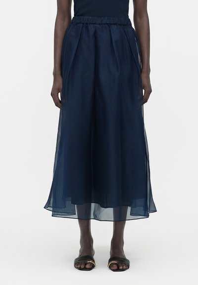 Model wearing navy blue sheer wide-leg pants with elastic waistband and black open-toe sandals against plain background.