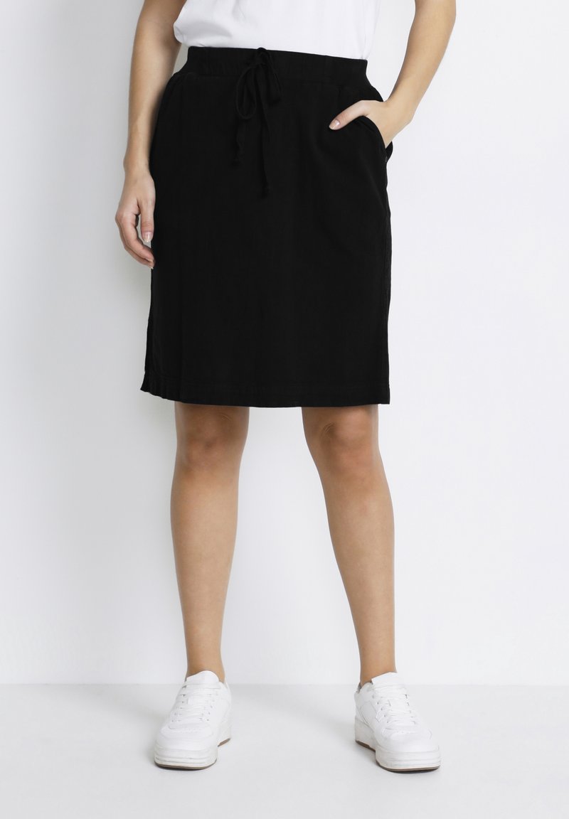 Femme portant une jupe noire mi-longue avec taille à cordon et poches, associée à des baskets blanches et un haut blanc.