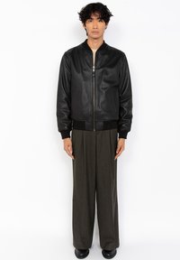 Hombre de pie mirando hacia adelante, vistiendo una chaqueta de cuero negra con cremallera, pantalones oscuros de pierna ancha y zapatos negros, contra un fondo blanco.