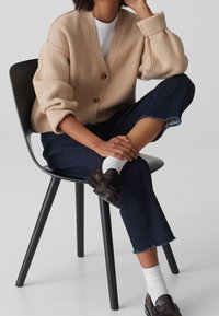 Cardigan beige a coste con scollo a V profondo, caratterizzato da tre bottoni, indossato sopra una t-shirt bianca. Abbinato a jeans scuri e mocassini marroni.