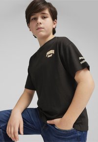 T-shirt di cotone nero con logo "Puma Sports" crema sul petto e logo Puma bianco sulla manica. Indossato con jeans blu.