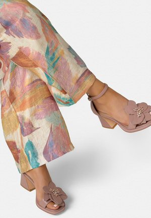Benenden met brede, kleurrijke, met patronen bedruktte broek en mauve sandalen met blokhakken, grote bloemversieringen en enkelbandjes.