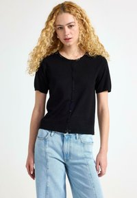 Černý krátký rukávový cardigan vyrobený z pleteného materiálu s kulatým výstřihem a zapínáním na knoflíky. Kombinovaný se světle modrými džíny.