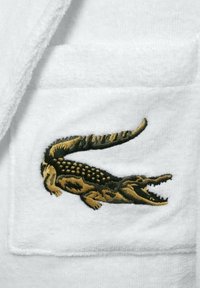 Un accappatoio bianco in tessuto con finitura testurizzata presenta un distintivo coccodrillo ricamato in nero e oro sulla tasca sinistra del petto.
