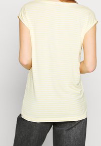 Žluté a bílé pruhované tričko vyrobené z měkké tkaniny, s krátkými rukávy, kulatým výstřihem a volným střihem s mírně zakřiveným lemem.