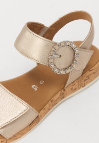 Gold synthetic leather sandal with ankle strap, circular chain accent, cork-textured wedge sole, and smooth tan footbed.