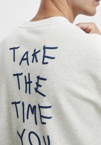 Jasnoszara bluza z haftowanym tekstem w granatowym kolorze na plecach, który mówi: "POŚWIĘĆ CZAS SOBIE." Okrągły dekolt i miękka tekstura.