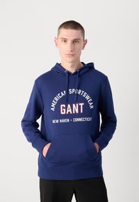Sudadera de color azul marino hecha de una mezcla de algodón, con un bolsillo canguro, capucha con cordón y texto gráfico en rojo y blanco en el pecho.