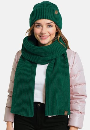 Ensemble bonnet et écharpe en tricot côtelé vert, fabriqué à partir de matériau doux. L'écharpe se drape autour du cou et présente une petite étiquette de logo.