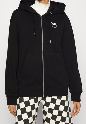Personne portant un sweat à capuche noir zippé avec un petit logo d'oiseau blanc et un pantalon à carreaux noir et blanc.