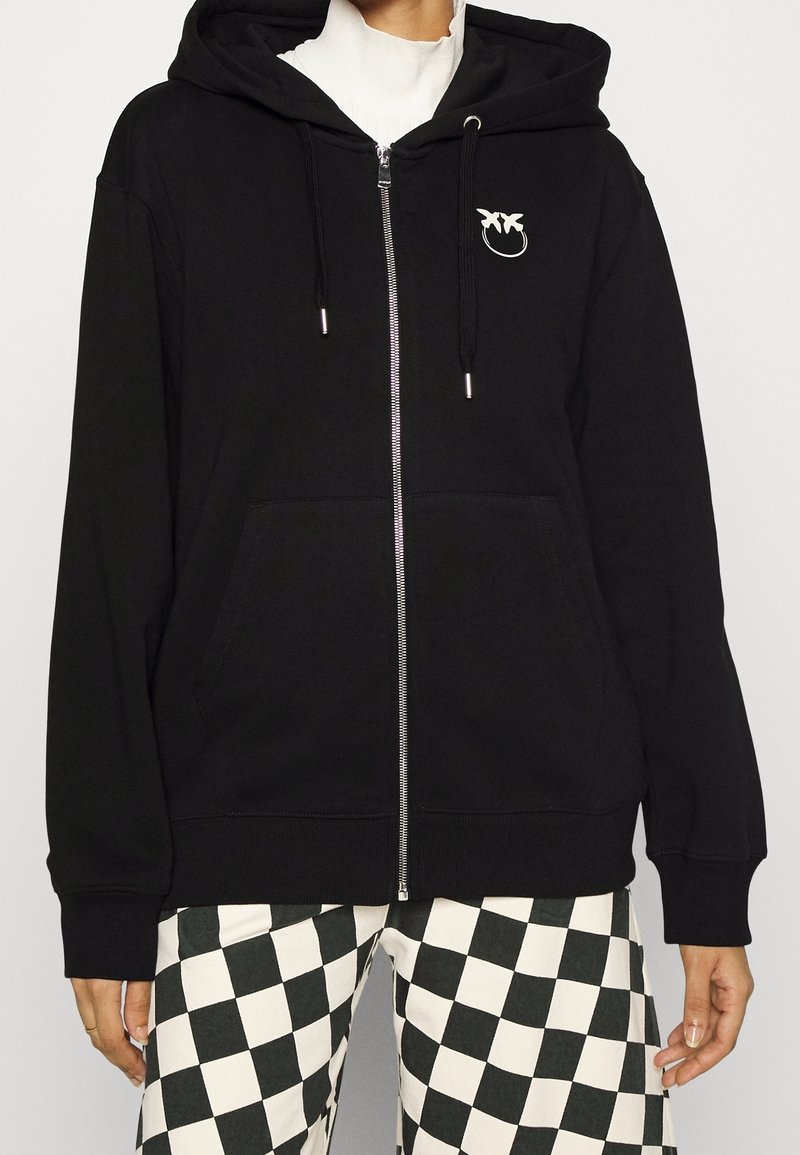 Personne portant un sweat à capuche noir zippé avec un petit logo d'oiseau blanc et un pantalon à carreaux noir et blanc.