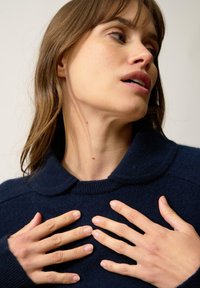 Granatowy sweter z okrągłym dekoltem, prążkowanymi mankietami i gładką teksturą. Ręce modelki delikatnie spoczywają na jej klatce piersiowej, podkreślając szczegóły.