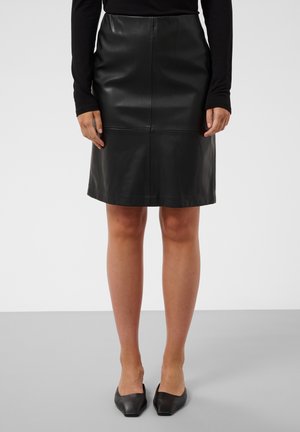 Femme portant une jupe en cuir noire longueur genou, un haut à manches longues noir et des chaussures plates noires à bout pointu, debout sur un sol gris.