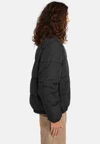 Person med krøllet brunt hår iført en sort pufferjakke og beige bukser, som står i profil og vender mod højre på en hvid baggrund.