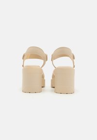 Beige Plateausandalen mit einem klobigen Absatz, verstellbarem Knöchelsriemen, glatter Textur und dezenten Stichdetails an den Riemen.