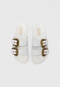 White patent slide sandals with two adjustable straps featuring ornate gold-tone buckles and a padded footbed.