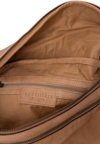 Interno di una borsa in pelle marrone con una tasca con zip, foderato in tessuto e un'etichetta che recita "RE:DESIGNED EST. 2003". Texture liscia, dettagli minimi.