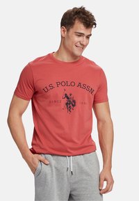 Crvena pamučna majica s okruglim izrezom, s mornarskoplavim grafičkim prikazom igrača polo i natpisom "U.S. POLO ASSN. SINCE 1890" jasno istaknutim.