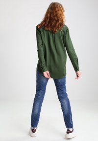 Camicia verde a maniche lunghe in tessuto liscio, con polsini con bottone e un accento arancione sulla spalla, abbinata a jeans blu.