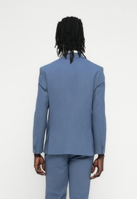 Ανοιχτό μπλε σακάκι με ραμμένη σχεδίαση, λοξές πέτες και κλείσιμο με ένα κουμπί. Λείο ύφασμα με ορατές ραφές και δύο ανοίγματα στο πίσω μέρος.