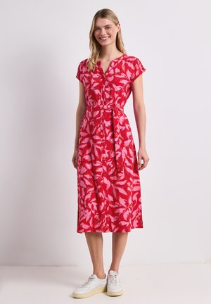 Frau lächelt, trägt ein rotes und pinkes geblümtes midikleid mit Knöpfen, kurzen Ärmeln und weißen Sneakern, steht vor einem einfarbigen hellen Hintergrund.