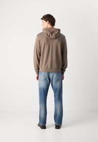 Sudadera con capucha marrón, con puños y dobladillo acanalados, combinada con jeans vaqueros claros y desgastados. Ambos artículos tienen un diseño casual y de corte relajado.