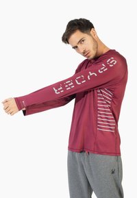 Bordeaux atletisk langærmet skjorte med et reflekterende logo på ærmet og stribet mønster på siden, parret med grå sweatpants.