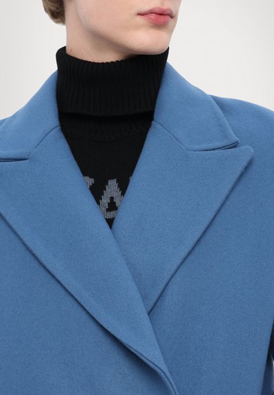 Svetlomodrý kabát z vlnenej zmesi s výstrihom do "V" je preložený cez čierny rebrovaný rolák so šedým geometrickým textovým dizajnom.