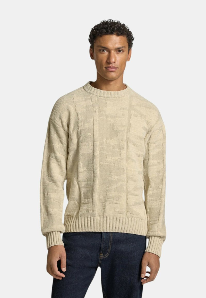Beige stickad tröja med strukturerad mönster, rund halsringning och ribbade ärmslut och nederkant. Bärs tillsammans med mörkblå jeans.