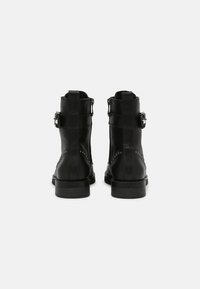 Černé kožené kotníkové boty s přezkami, texturovaným povrchem a nízkým hromadným podpatkem. Na zadní straně se nacházejí dvojité pásky a dekorační hroty.
