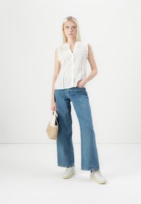 Weißes, ärmelloses, knöpfbares Blusenshirt aus strukturiertem Stoff mit Spitzenbesatz, kombiniert mit hochgeschnittenen blauen Jeans und weißen Sneakers. Hält eine kleine Tasche.