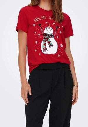 Vrouw draagt een rood T-shirt met een glimlachende sneeuwmanafbeelding en tekst "Hug me, I’m", gecombineerd met een zwarte broek.