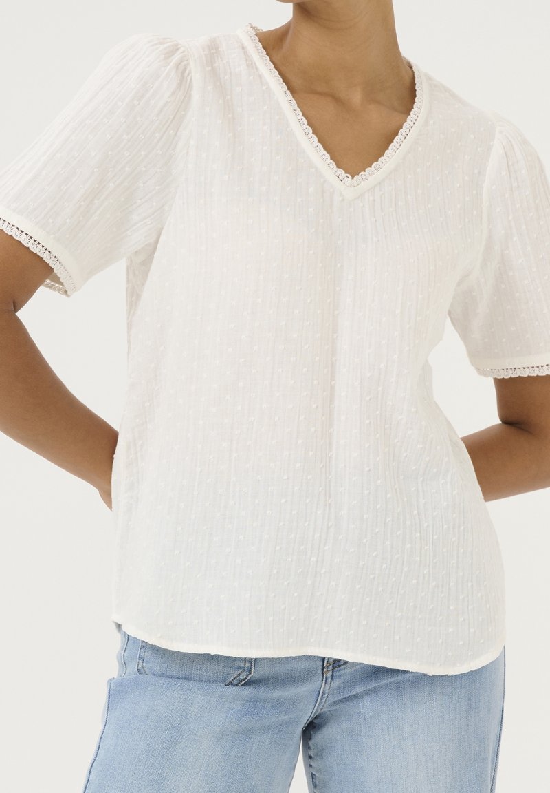 Camiseta blanca de manga corta con escote en V, fabricada con tela texturizada y con delicado borde de encaje en el cuello y las mangas. Corte informal, forma ligeramente holgada.