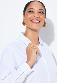 Witte blouse met een kraag en lange mouwen. Heeft een gladde textuur en een kleine knop bij de manchetten. Gouden creolen.